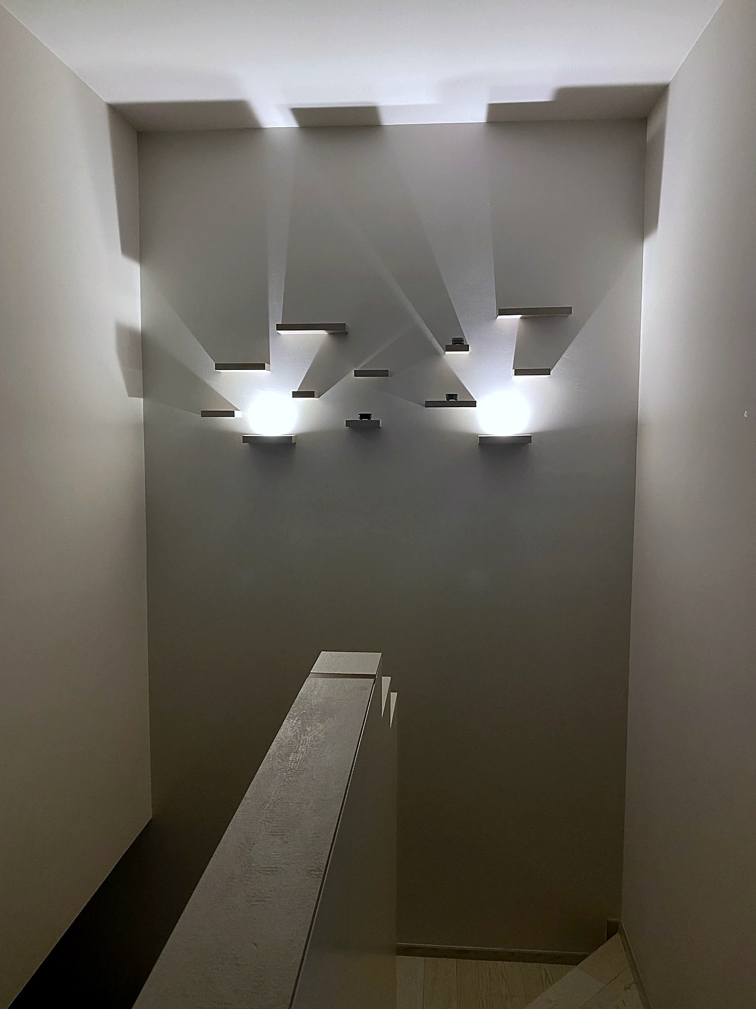 Vano scala: mensole e sorgenti a parete disegnano ombre geometriche, la parete diventa composizione.