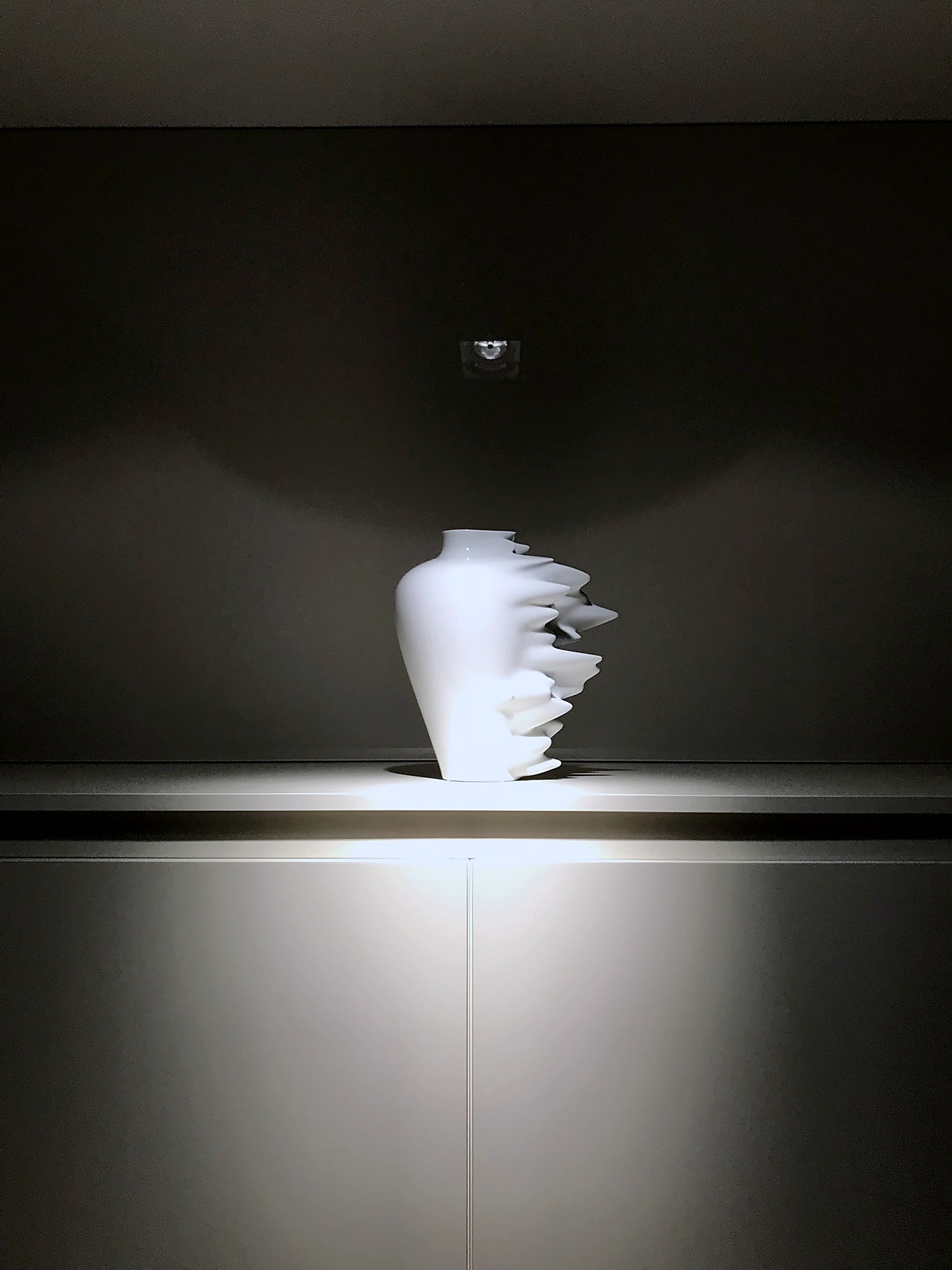 Luce d'accento dall'alto: l'oggetto emerge dal fondo scuro e la luce prosegue sulle ante.